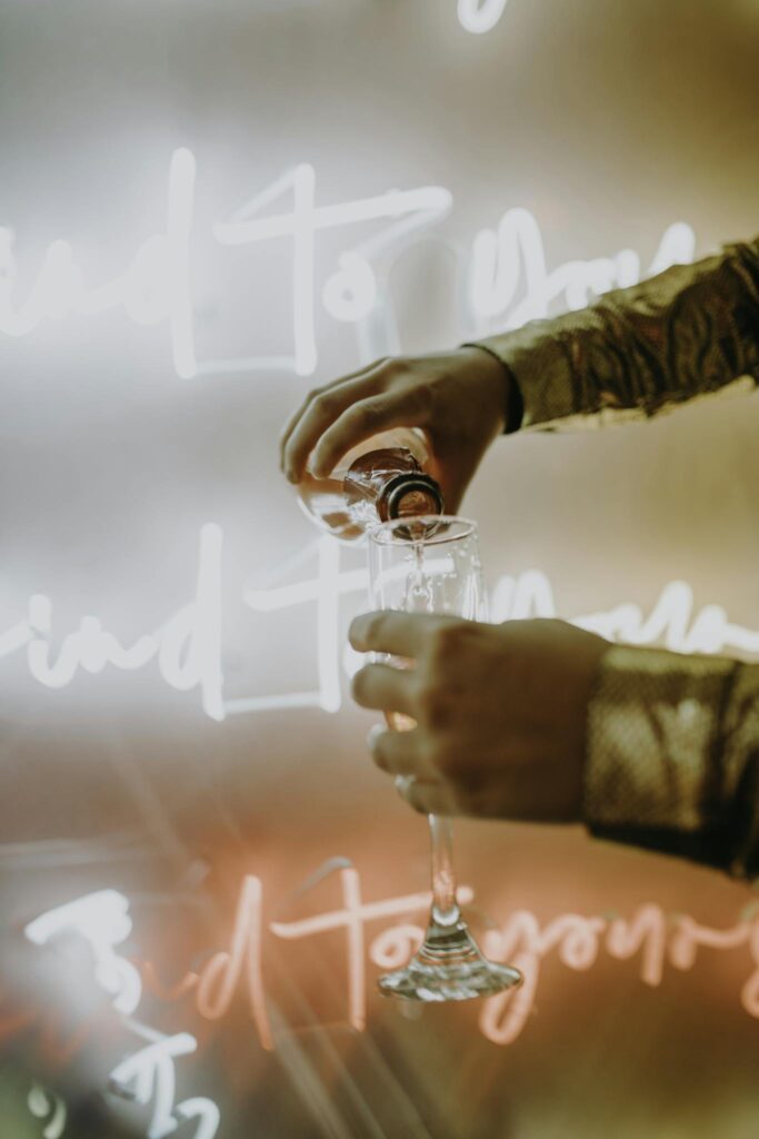 A close-up of hands pouring champagne against a festive neon-lit background.